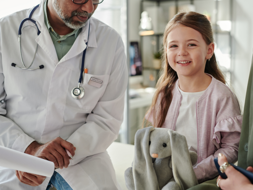 little girl is pictured sitting with a doctor in a specialist children's hospital holding a grey bunny teddy bear. The girl is smiling and wearing a pink top, she has brown hair.