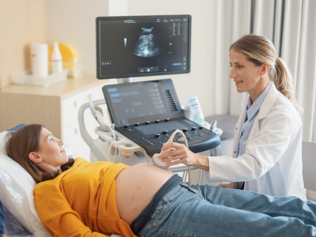 Pregnant woman having a scan with a female doctor. The pregnant white woman is wearing an orange jumper and blue jeans. The female white doctor is wearing a white lab coat and has blonde hair in a ponytail.